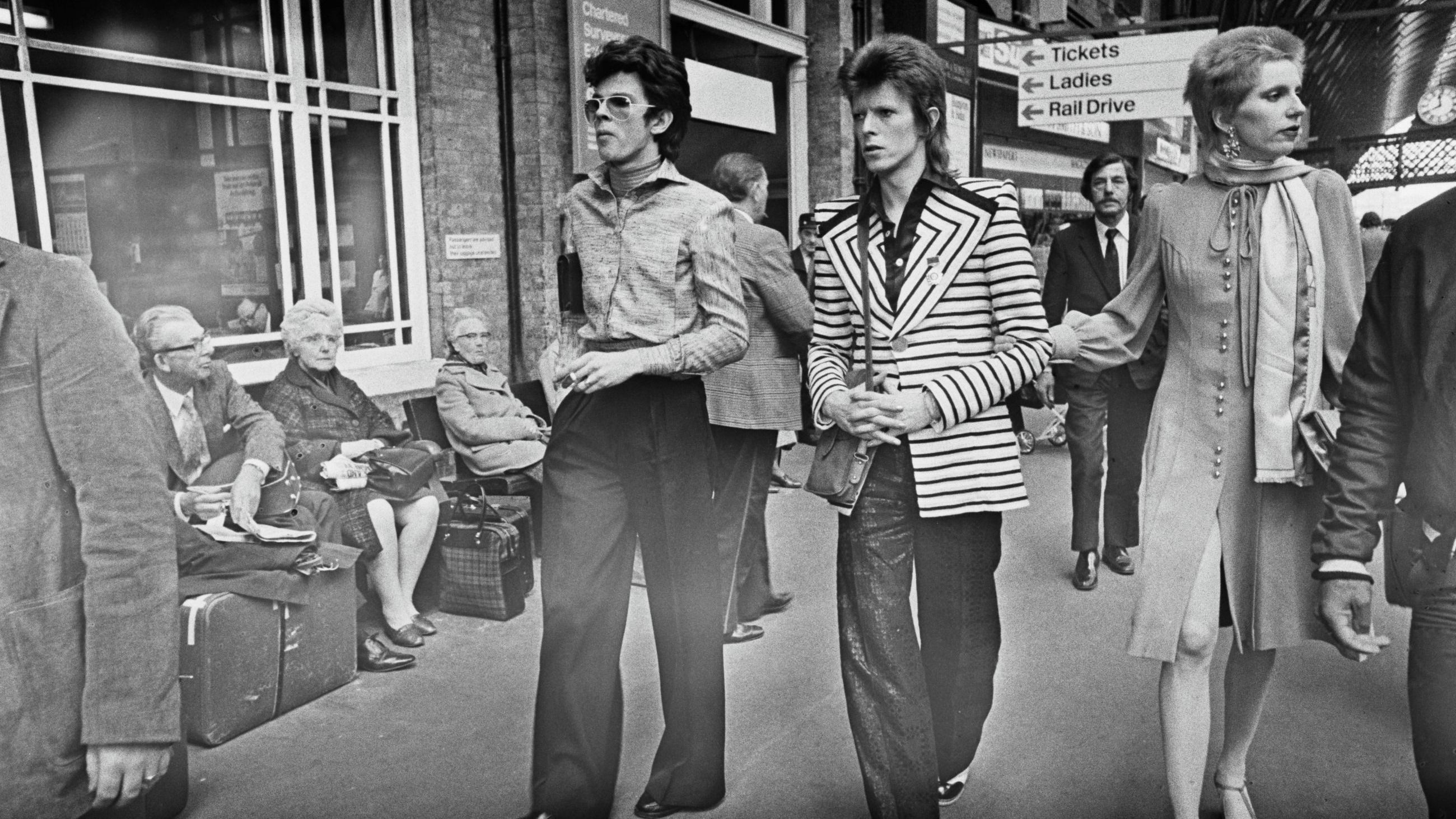 David Bowie alla stazione di King's Cross a Londra con sua moglie Angie Bowie e lo stilista inglese Freddie Burretti, mentre parte per Aberdeen in Scozia per il suo tour Ziggy Stardust, Regno Unito, 15 maggio 1973 - Credits Getty Images
