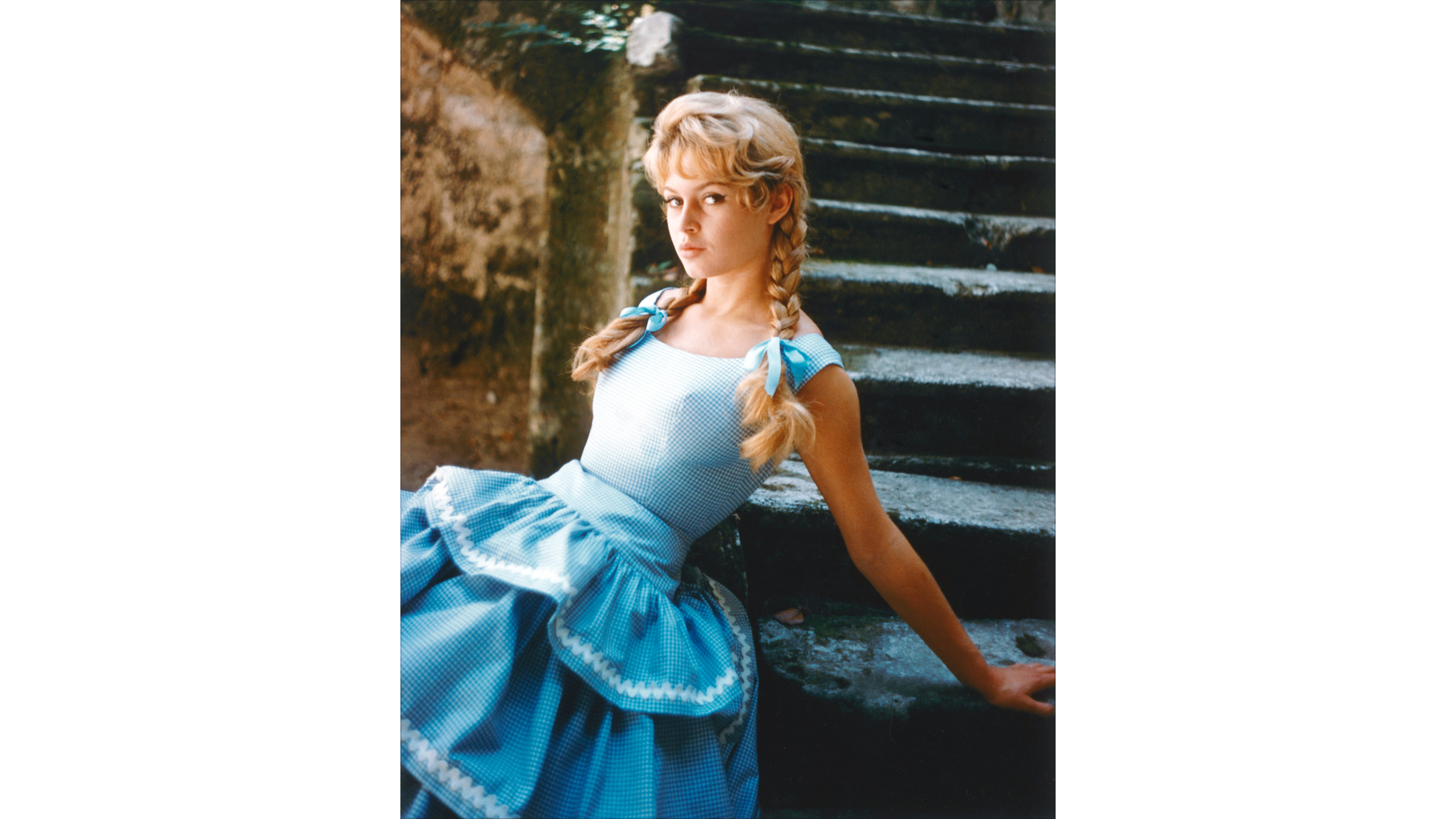 Brigitte Bardot indossa un abito a quadretti blu con i capelli intrecciati in trecce, legate con fiocchi blu, seduta su una serie di gradini in pietra, nel 1960 - Credits: Getty 