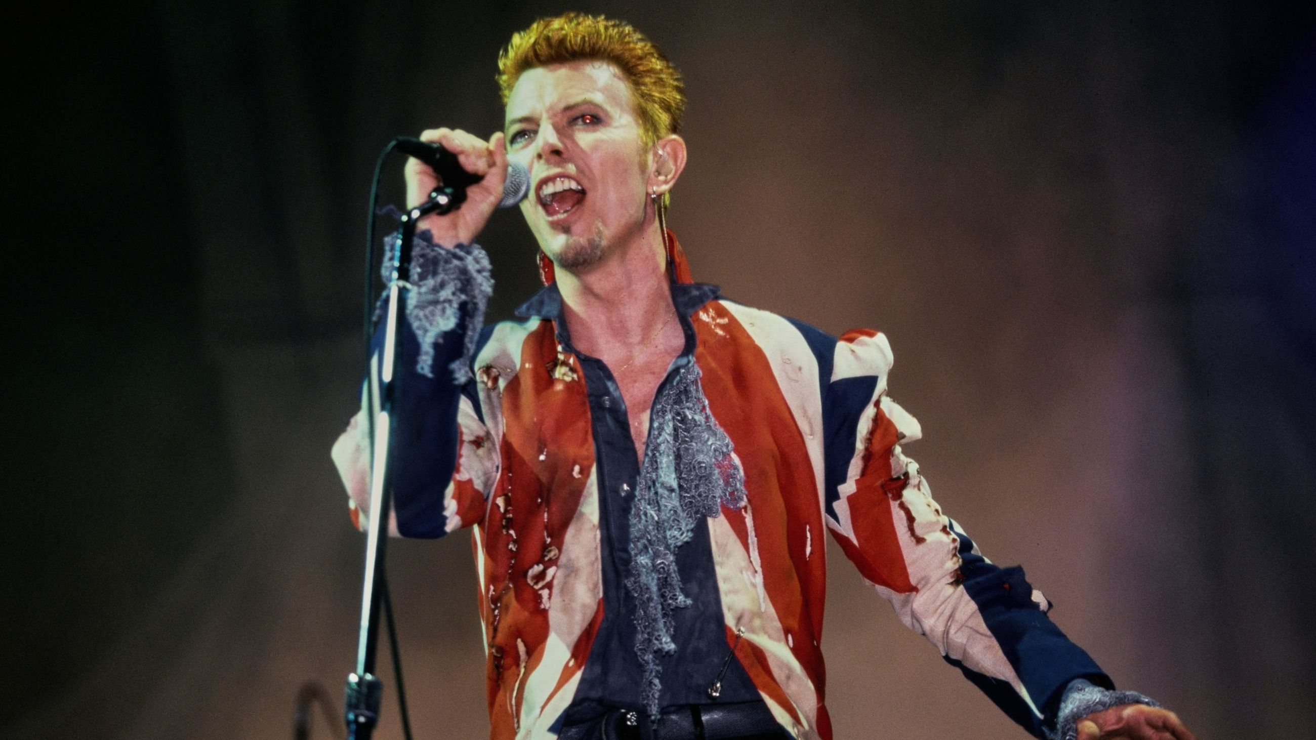 David Bowie, con indosso la sua “Union Jacket”, si esibisce dal vivo al Phoenix Festival, presso il Long Marston Airfield a Long Marston, Warwickshire, 1996. La giacca, caratterizzata dal disegno della bandiera britannica, è stata creata da Alexander McQueen - Credits Getty Images
