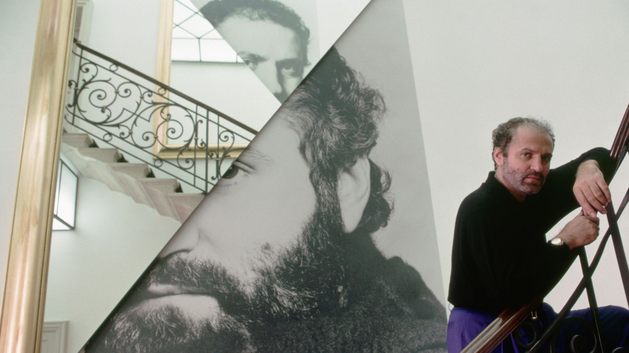 Lo stilista Gianni Versace posa sulle scale di casa sua. Sullo sfondo una composizione realizzata con alcune fotografie che ritraggono il designer; 1990 - Credits: Getty Images